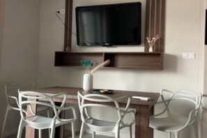 A dining area with a wooden table, white chairs, a flat-screen TV mounted above, and a decorative vase with a plant. - Half of a holiday home with a large garden (Slajszewo)