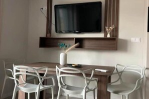 Un comedor con una mesa de madera, sillas blancas, un televisor de pantalla plana montado arriba y un pequeño estante con adornos.