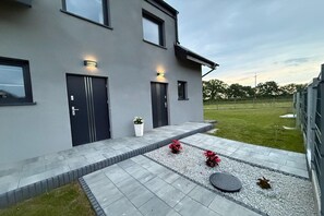 A modern two-story house with a grey exterior, two black doors, and a small garden area with gravel and potted plants. - Half of a holiday home with a large garden (Slajszewo)
