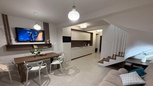 Un salon moderne comprenant un canapé, une table à manger avec des chaises, un téléviseur fixé au mur et un coin cuisine avec des placards et un four à micro-ondes.