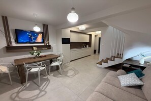 Una sala de estar moderna con un sofá, una mesa de comedor con sillas, un televisor montado en la pared y una zona de cocina con armarios y un microondas.