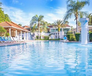Piscina com espreguiçadeiras, palmeiras e um edifício com telhado em azulejo.