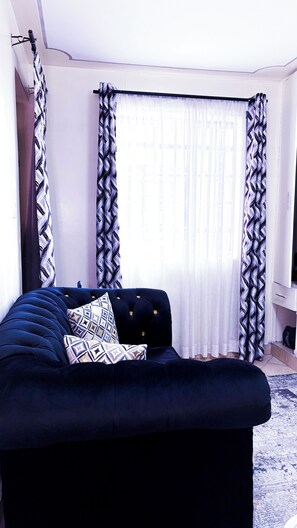A room with a dark blue sofa, patterned pillows, and sheer curtains with a geometric pattern. - Studio in south b near airport (Nairobi)