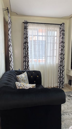 A living room with a black sofa, patterned cushions, and sheer curtains. - Studio in south b near airport (Nairobi)