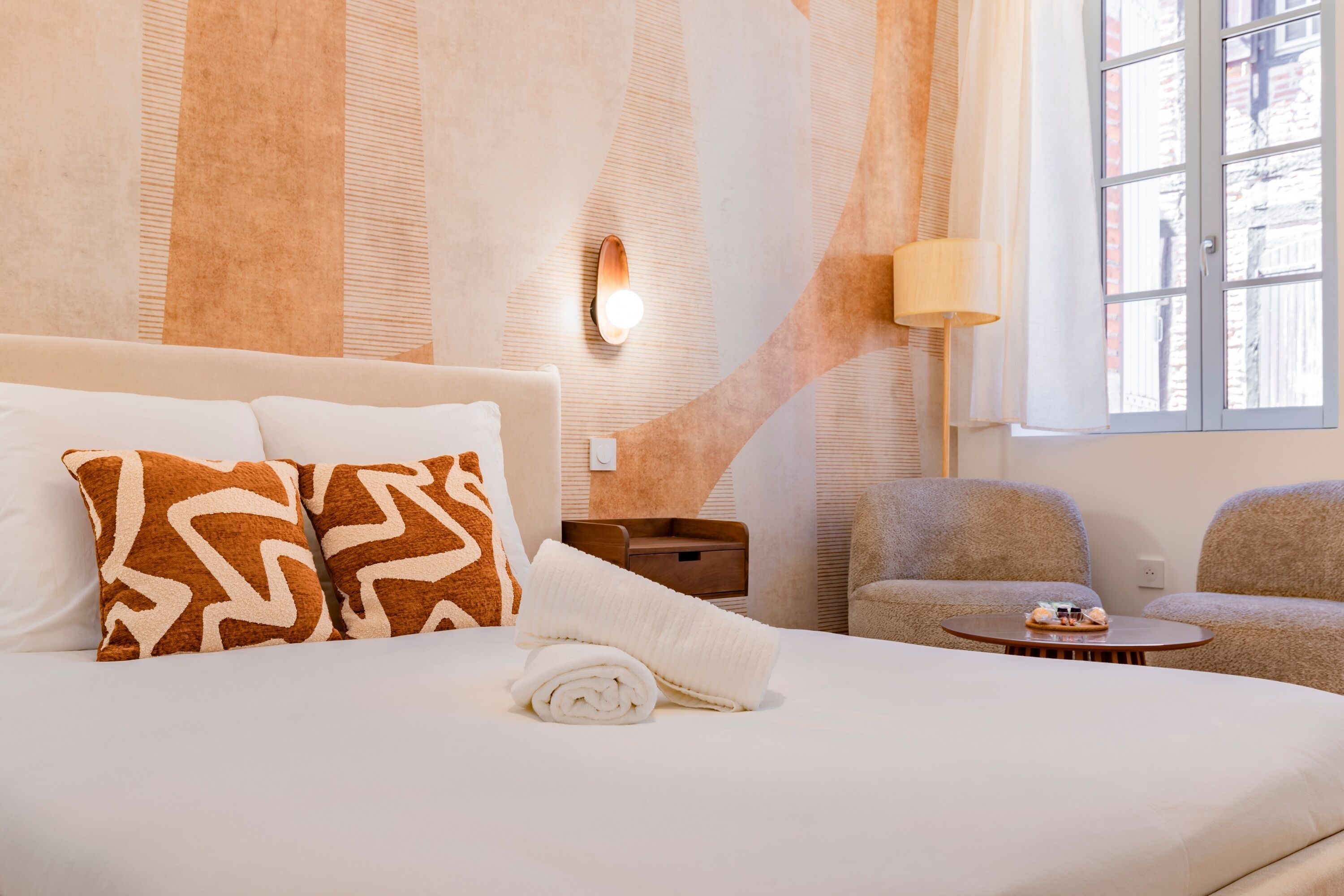A hotel room with a bed, two armchairs, a small table, a lamp, and a window with a view of buildings.