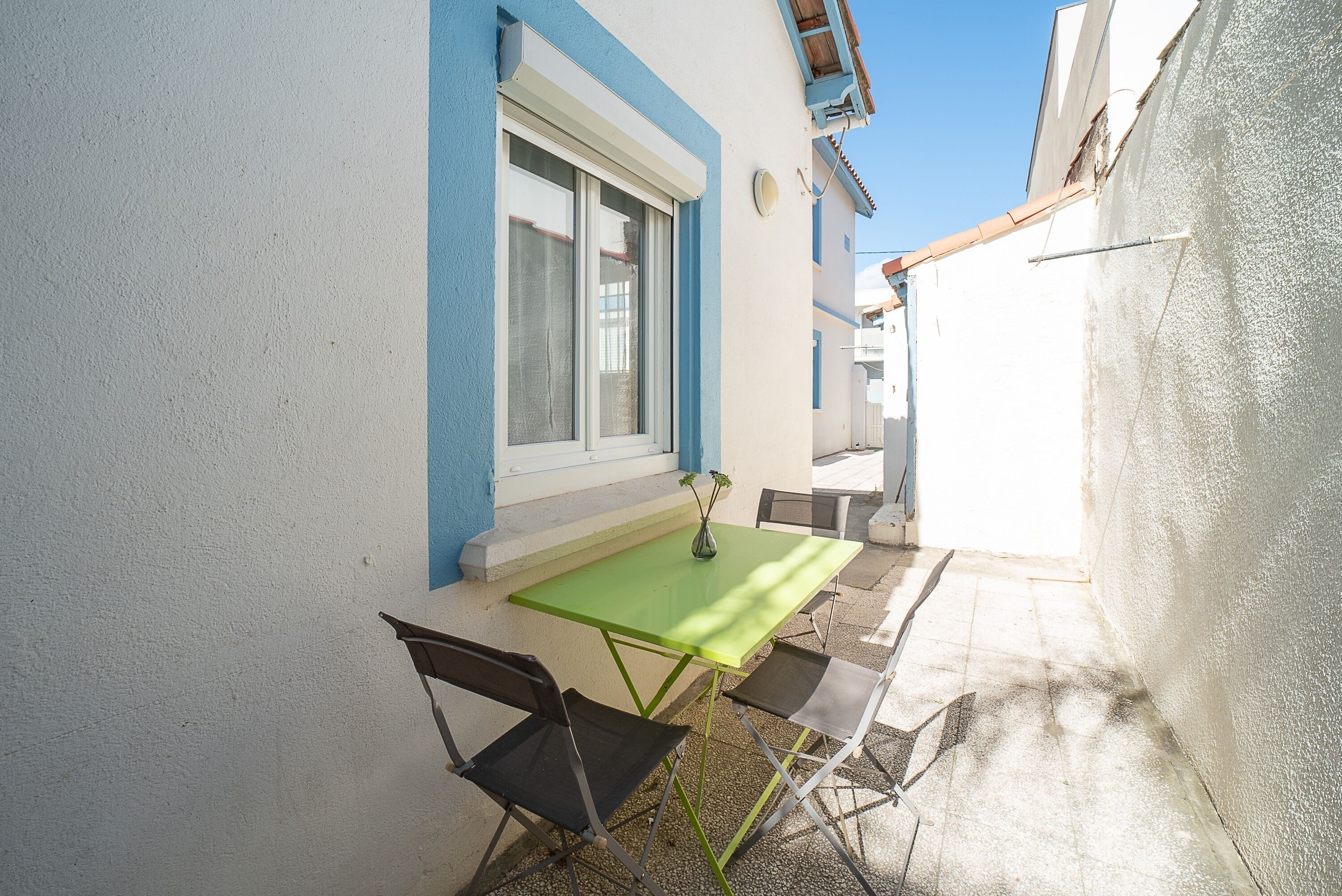 Une petite terrasse extérieure avec une table et des chaises, une fenêtre avec un rideau et un mur bleu et blanc.