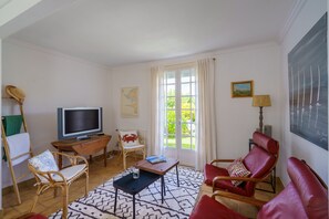 A living room with a television, wicker chairs, a red sofa, a patterned rug, and a large window with curtains. - Ty Cos-Ker - Dans le bourg de Crozon (Crozon)