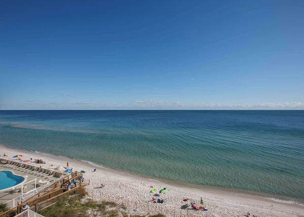 Ein Strand mit klarem, blauem Wasser, einem sandigen Ufer und einem Poolbereich mit Liegestühlen und Sonnenschirmen.