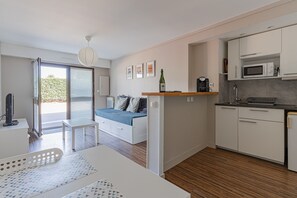 A modern kitchen with white cabinets, a microwave, and a coffee maker. There is a dining area with a table and chairs, and a bed with a blue blanket. - La Pause courseullaise - Joli studio proche plage (Courseulles-sur-Mer)