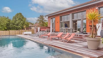 Une piscine avec des chaises longues, une terrasse en bois et un bâtiment doté de grandes fenêtres.