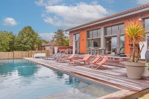 Ein Schwimmbecken mit Liegestühlen, einer Holzterrasse und einem Gebäude mit großen Fenstern.