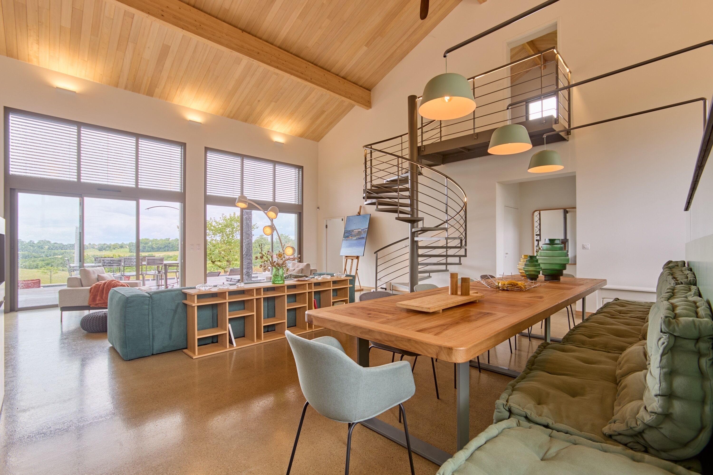Un vaste espace de vie comprenant une table à manger en bois, un escalier en colimaçon et de grandes fenêtres donnant sur l’extérieur.