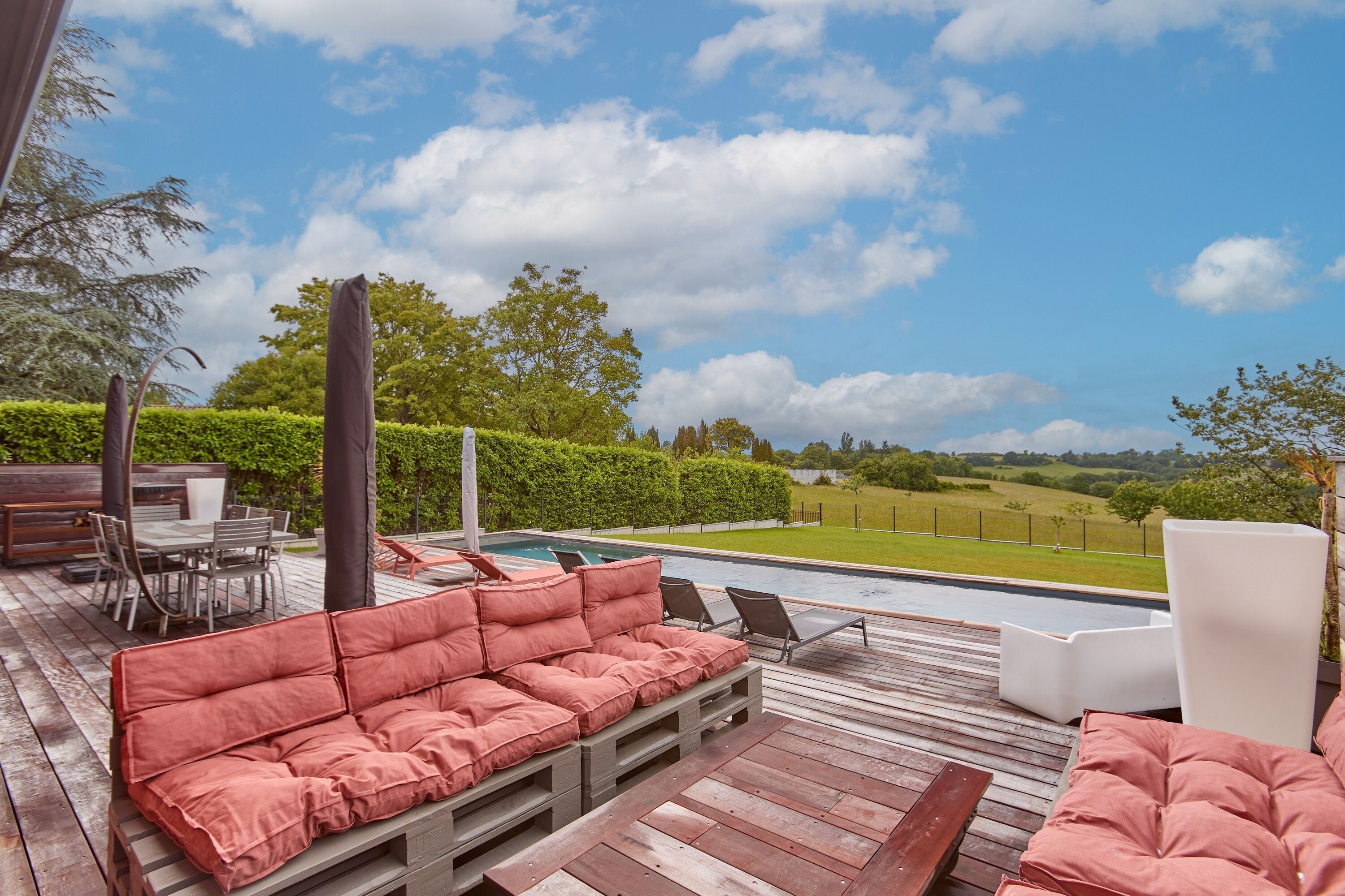 Une terrasse en bois avec un canapé rouge doté de coussins, une piscine et une vue sur un paysage verdoyant.