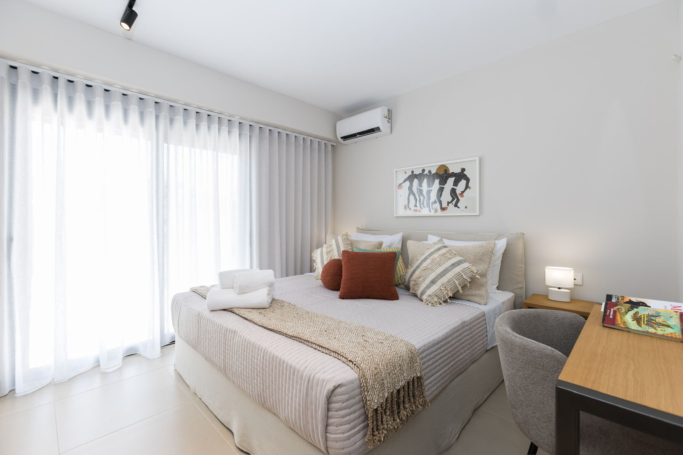 A modern hotel room with a bed, a desk, a chair, a window with curtains, an air conditioner, and a wall-mounted artwork.