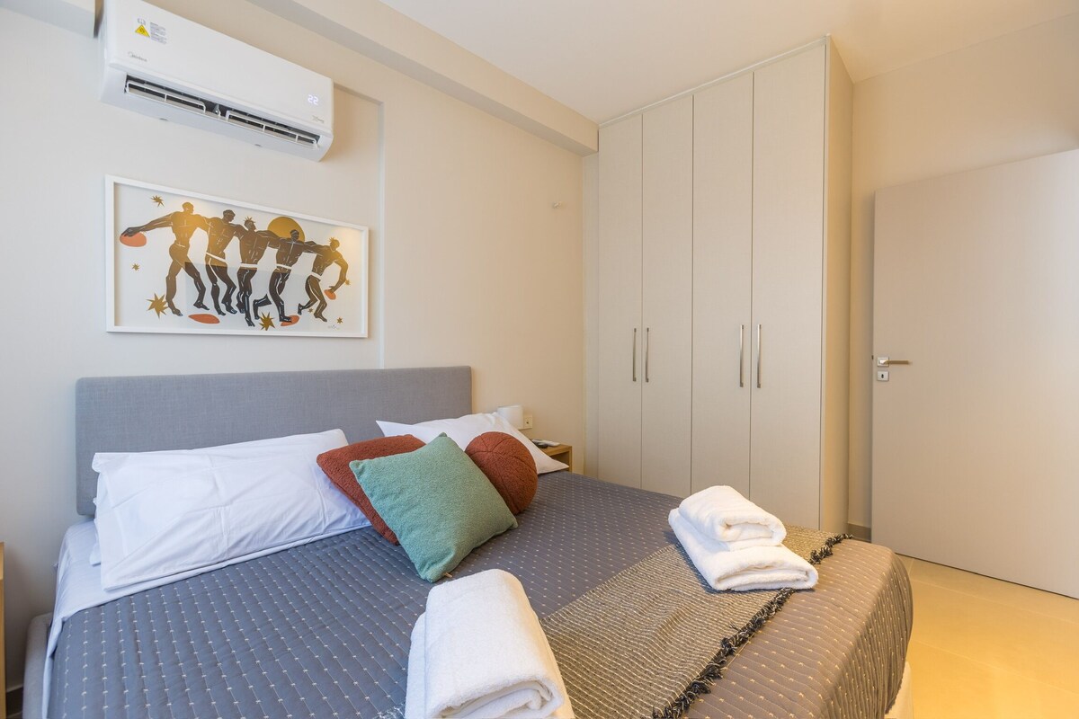 A hotel room with a bed, pillows, a wall-mounted air conditioner, a framed artwork, and a wardrobe.