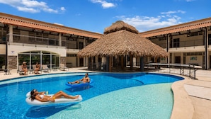 A swimming pool with two people on inflatable floats, a thatched-roof structure, and a building with balconies.