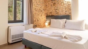 A bedroom with a stone wall, a bed with white linens, a wooden ceiling, and a window with curtains. - Villa Elpida (Chania)