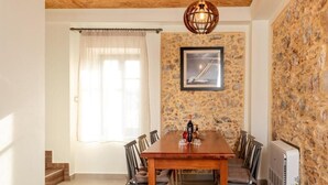 A dining area with a wooden table, chairs, a framed picture on the wall, a window with curtains, and a stone wall. - Villa Elpida (Chania)