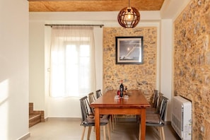 A dining area with a wooden table, chairs, a framed picture on the wall, a window with curtains, and a stone wall. - Villa Elpida (Chania)