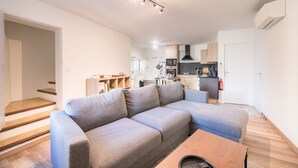 A modern living room with a grey sectional sofa, wooden flooring, a kitchen in the background, and a staircase leading to another level. - La Sacristie - Au coeur du village de Donzère (Donzère)