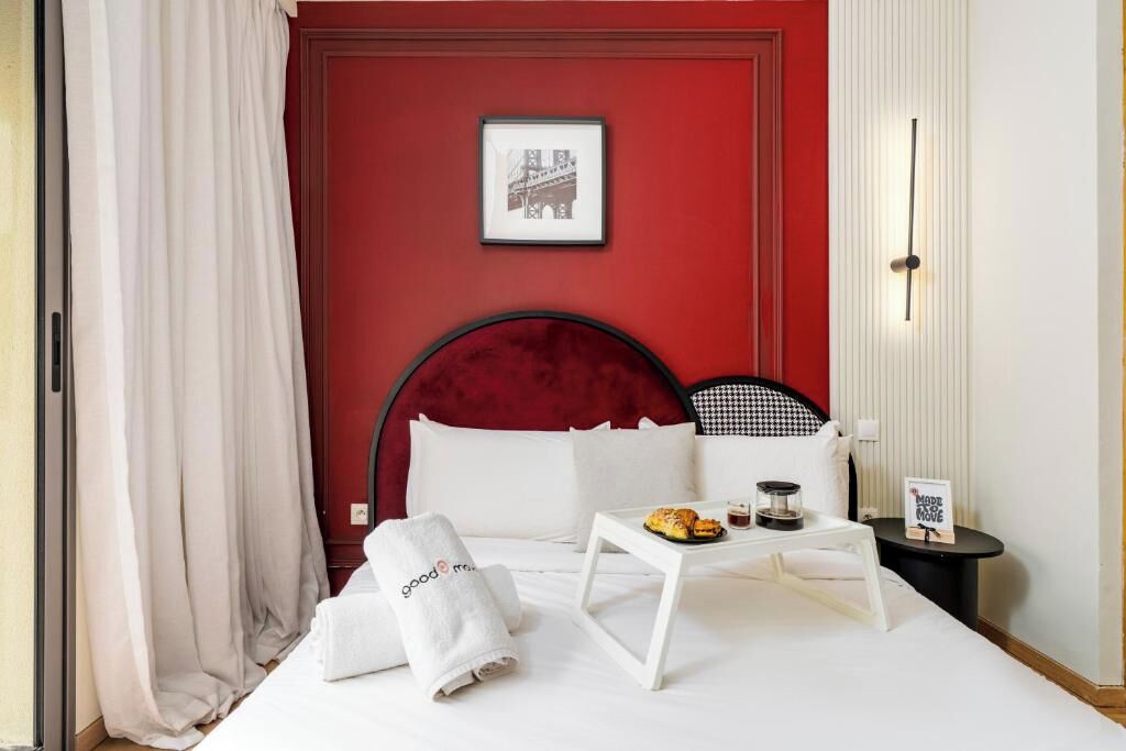 Camera d'albergo con parete di accento rossa, letto con lenzuola bianche, un quadro incorniciato, vassoio con pasticcini, un asciugamano e un piccolo tavolo.