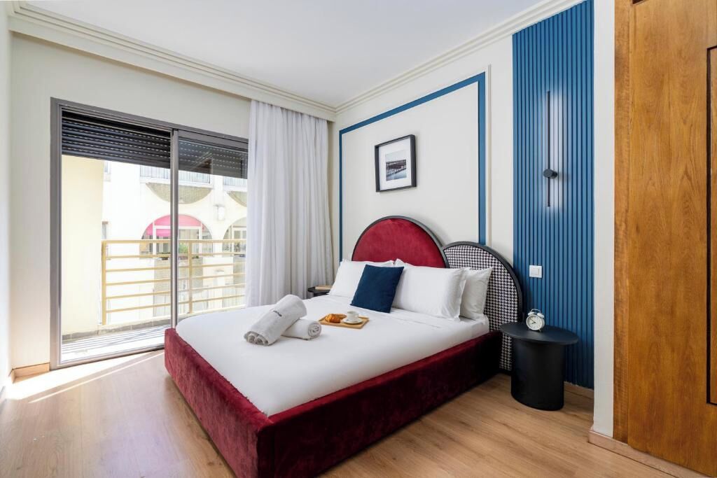 Ein Hotelzimmer mit einem Bett, einem Nachttisch, einer Uhr und Blick ins Freie.