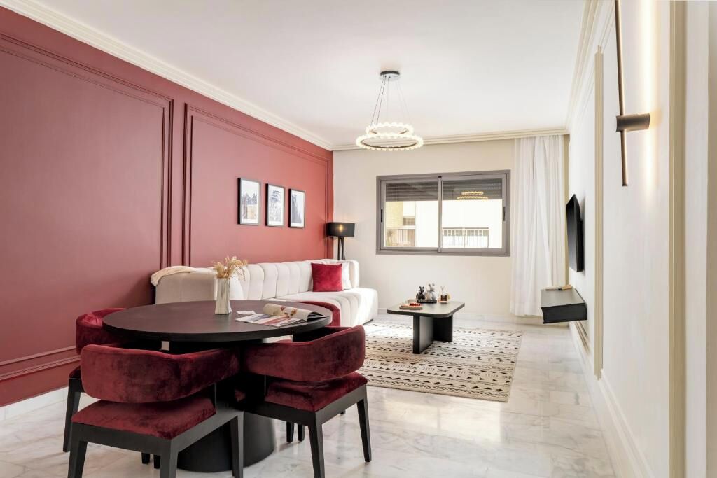 Uma sala de estar moderna com uma mesa de jantar redonda, cadeiras estofadas em vermelho, um sofá branco e uma janela grande com cortinas.