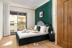 Habitación de hotel con cama, mesita de noche, armario, ventana con vistas a edificios y balcón.