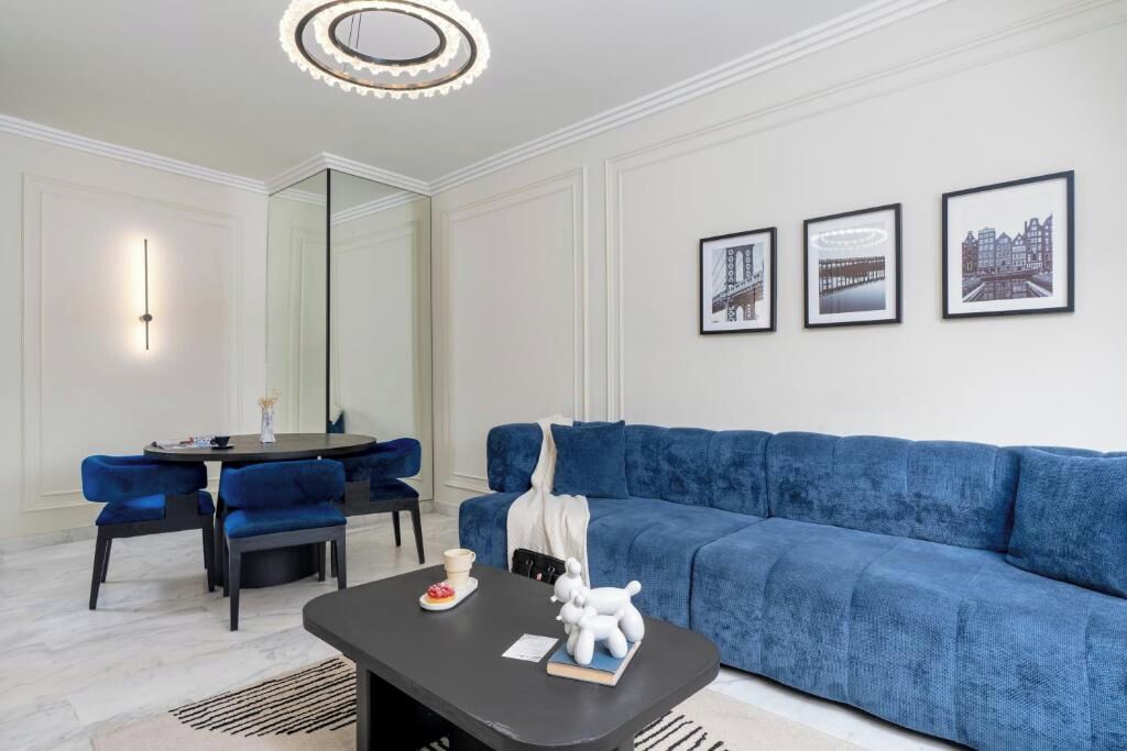 Un salon moderne avec un canapé bleu, une table basse noire ornée d’une figurine de chien, un coin repas avec des chaises bleues et, au mur, des tableaux encadrés représentant des paysages urbains.