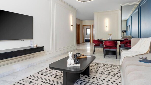 Ein modernes Wohnzimmer mit einem Flachbildfernseher, einem schwarzen Couchtisch mit Dekoration, einem gemusterten Teppich, einem roten Sofa und einem Essbereich mit roten Stühlen.