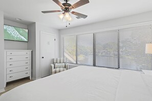 Ein Schlafzimmer mit einem Bett, einem Deckenventilator, einem Fernseher, einem Schminktisch, einem Stuhl und großen Fenstern mit Jalousien.