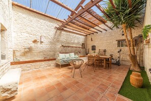 Ein Zimmer mit Steinwänden und Holzbalkendecke, ausgestattet mit einer Couch, einem Esstisch, Stühlen und einem kleinen Baum.