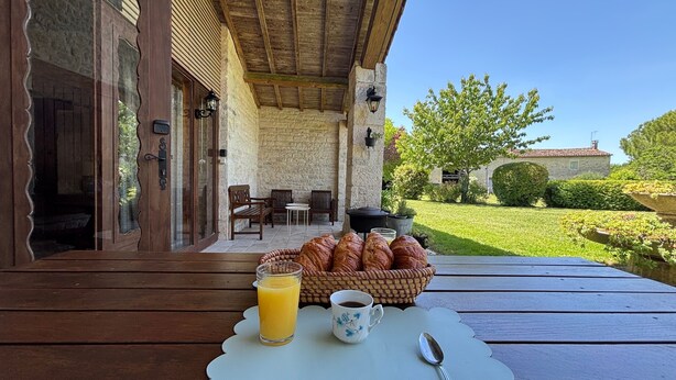 Ein Holztisch mit einem Korb voller Croissants, einem Glas Orangensaft und einer Tasse Kaffee, auf einer Terrasse mit Steinmauern und Sitzgelegenheiten.