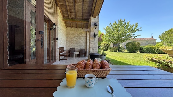 Une table en bois, sur laquelle sont posés un panier de croissants, un verre de jus d’orange et une tasse de café, est installée sur une terrasse aux murs de pierre, agrémentée de sièges d’extérieur.
