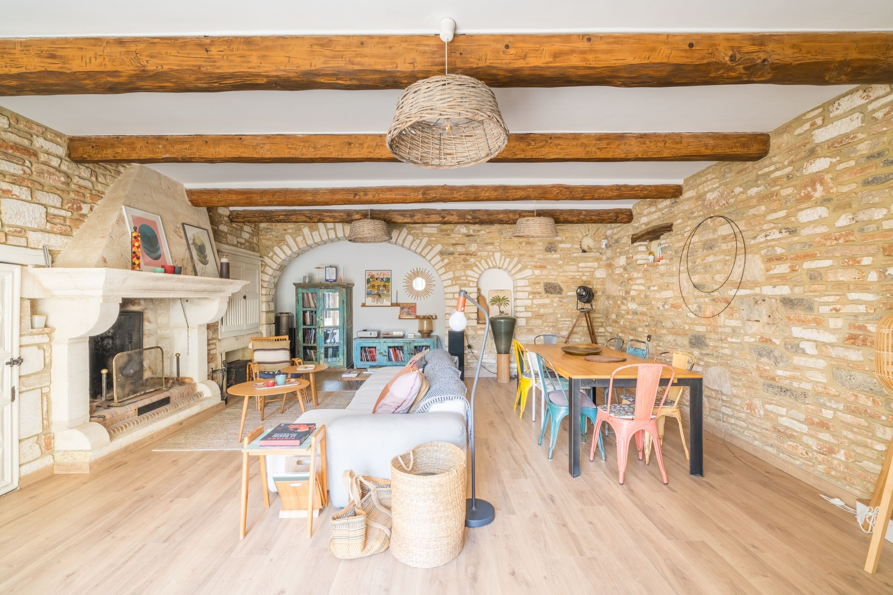 Un interior rústico con vigas de madera a la vista, pared de piedra, chimenea, sofá, mesa de comedor y sillas de colores.