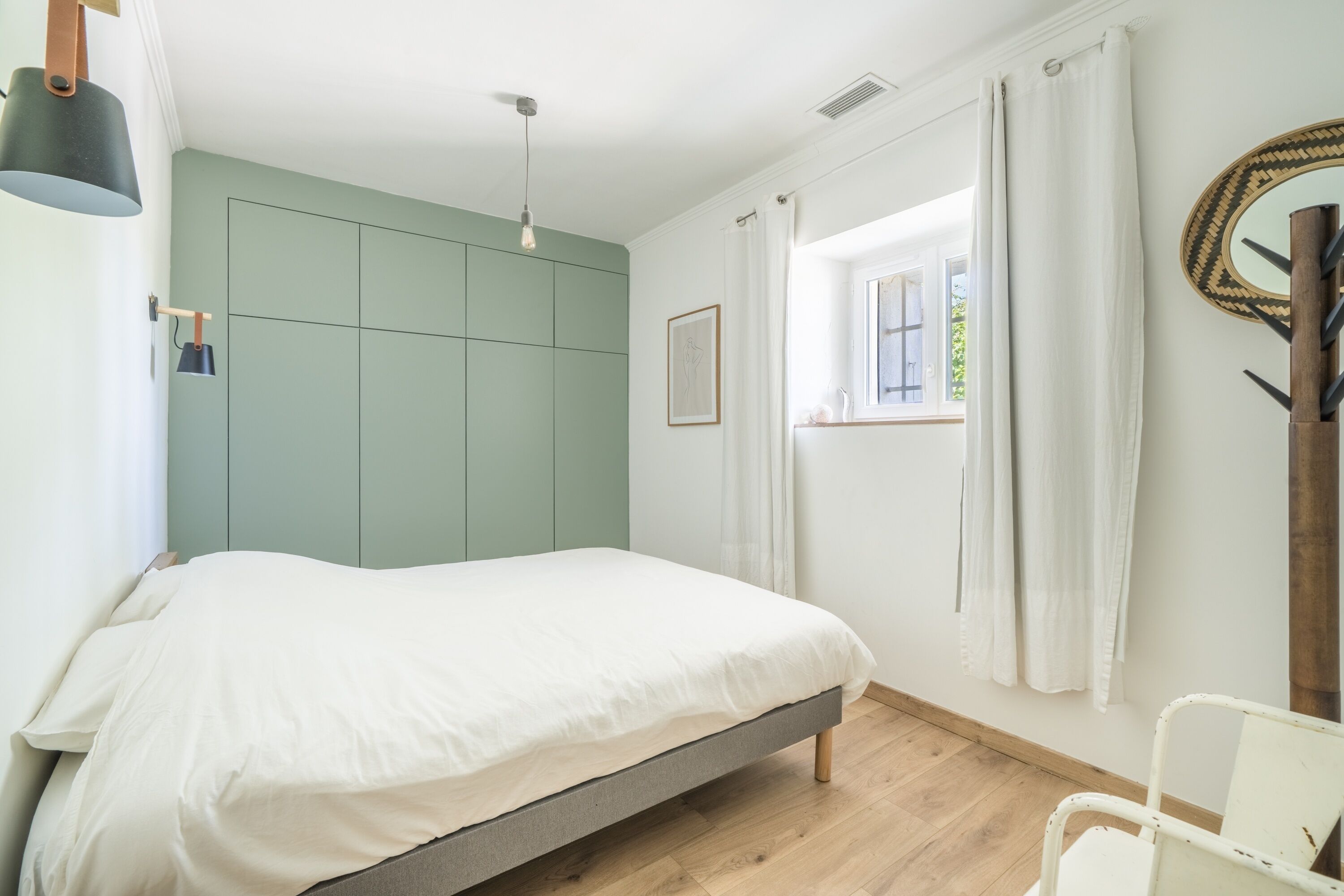 A bedroom with a bed, a window with curtains, a wall-mounted mirror, and a bedside lamp.