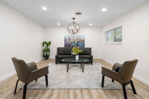 Una sala de estar moderna con un sofá, dos sillones, una mesa de centro, una araña, un cuadro, una planta y una ventana con persianas.