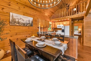 A wooden interior with a dining table set for six, a kitchen area, and a painting on the wall. - Wonder View Cabin! (Maggie Valley)