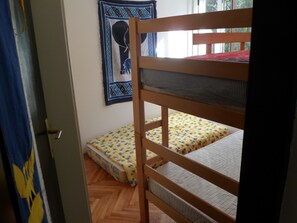 Ein Stockbett mit Matratze und Decke, eine Tür mit Griff, ein Wandbehang mit Muster und ein Fenster mit Tageslicht.
