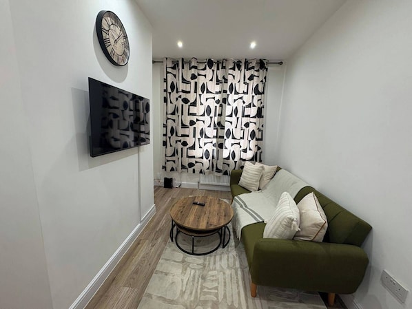 A modern living room with a green sofa, a round wooden coffee table, a wall clock, and abstract wall art.