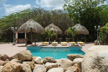 A pool area with thatched umbrellas, lounge chairs, and a small structure, surrounded by rocks and trees.
