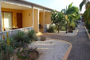Un hotel con una zona de estar exterior techada, un sendero de grava y diversas plantas.