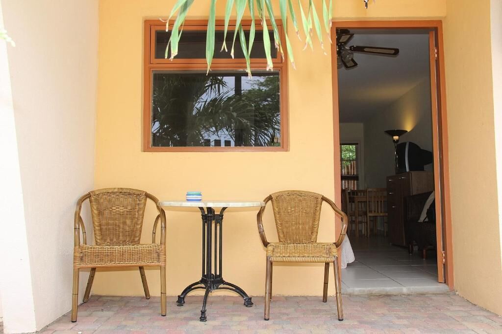 Una habitación con dos sillones de mimbre, una mesita, un ventanal con vistas a palmeras y una puerta que da a otra habitación.