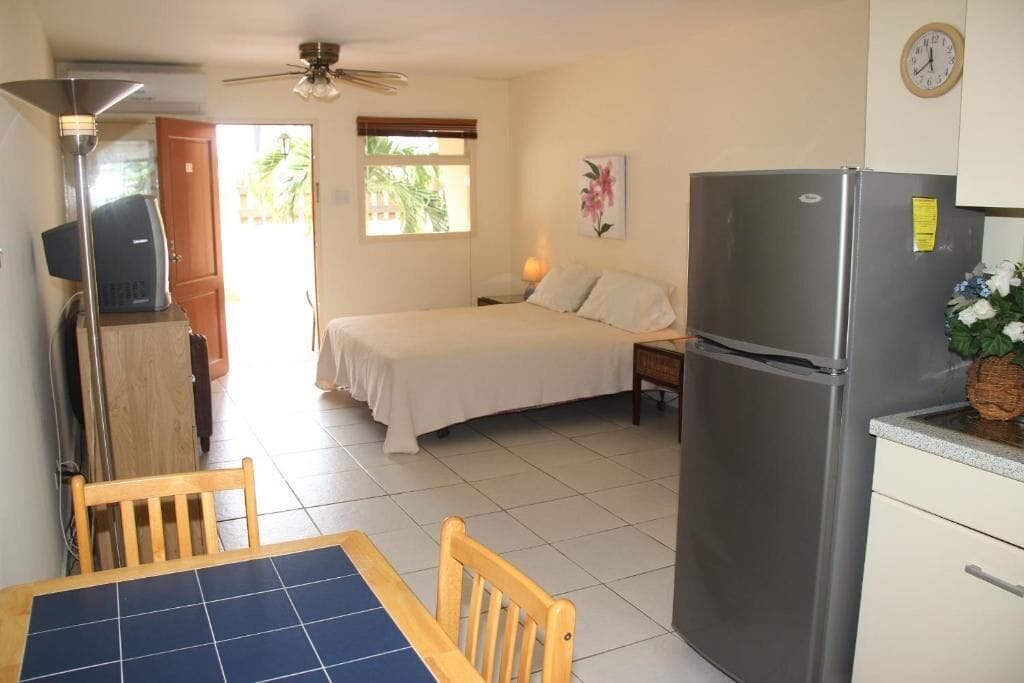 A compact room with a bed, a refrigerator, a dining table with chairs, a ceiling fan, a window with blinds, and a clock on the wall.