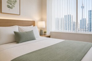Ein Hotelzimmer mit einem Bett, einem Nachttisch, einer Lampe, einem Fenster mit Jalousien und Blick auf die Stadt.