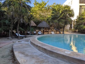 Una piscina con sillones de descanso, palmeras y una estructura de techo de paja.