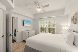 Een slaapkamer met een bed, een plafondventilator, een televisie, een raam met jaloezieën en een deur die leidt naar een andere kamer.