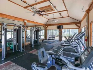 Un gimnasio con diversos equipos de ejercicio, un ventilador de techo y ventanas amplias.