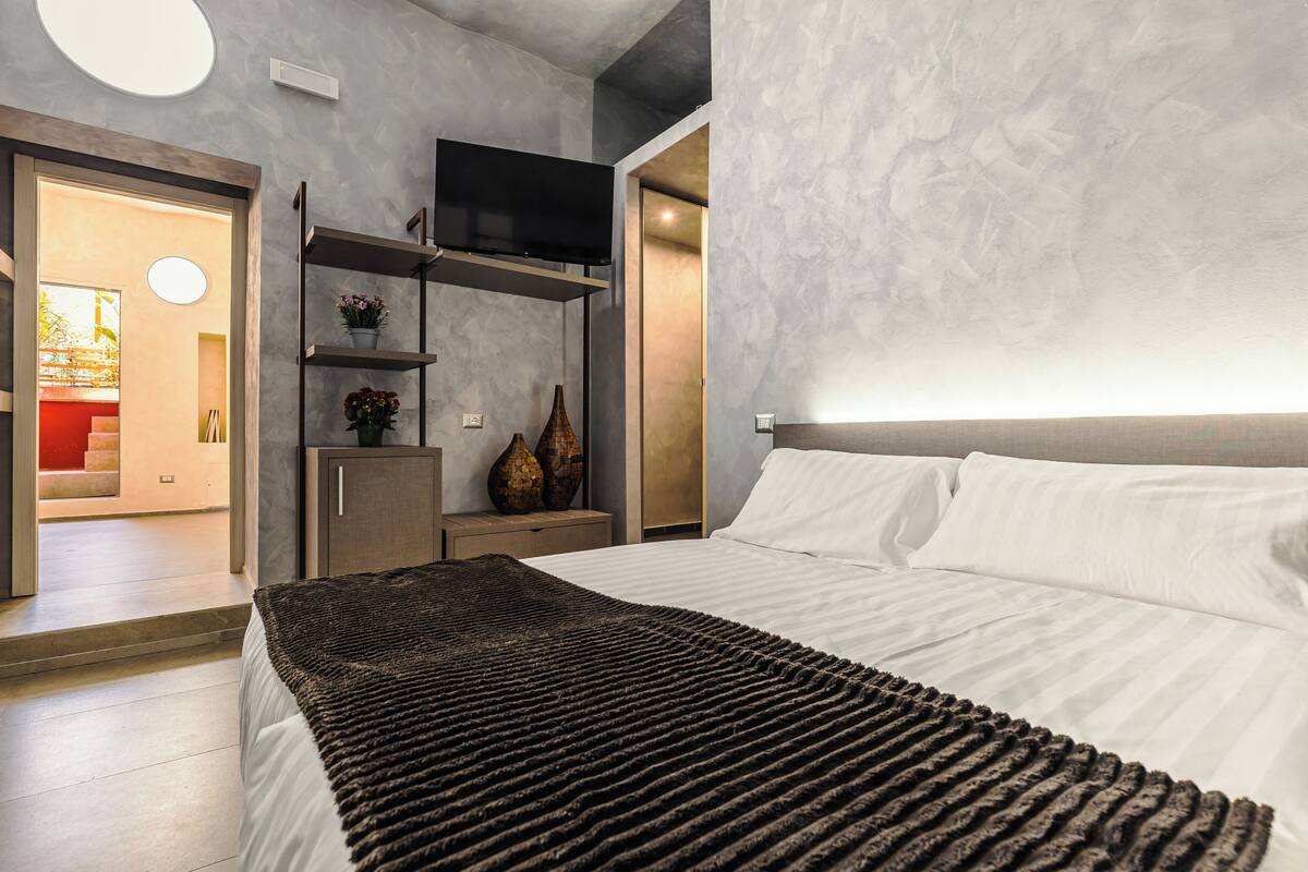 A modern bedroom with a bed, a TV, a shelf with decorative items, and a view of the outside.