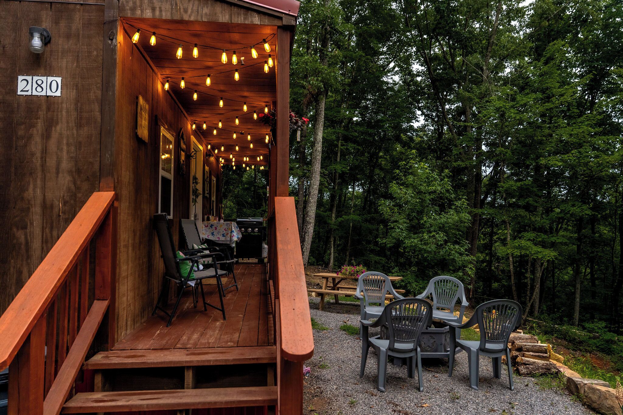 A wooden cabin with a porch, numbered 280, featuring outdoor seating with plastic chairs and a table, surrounded by trees.